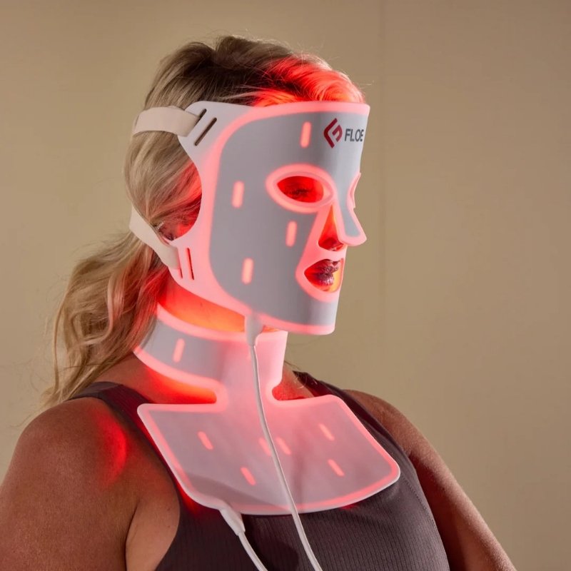A woman wearing a Floe LED light therapy mask on her face and neck, with the mask glowing red and pink against a plain beige background.