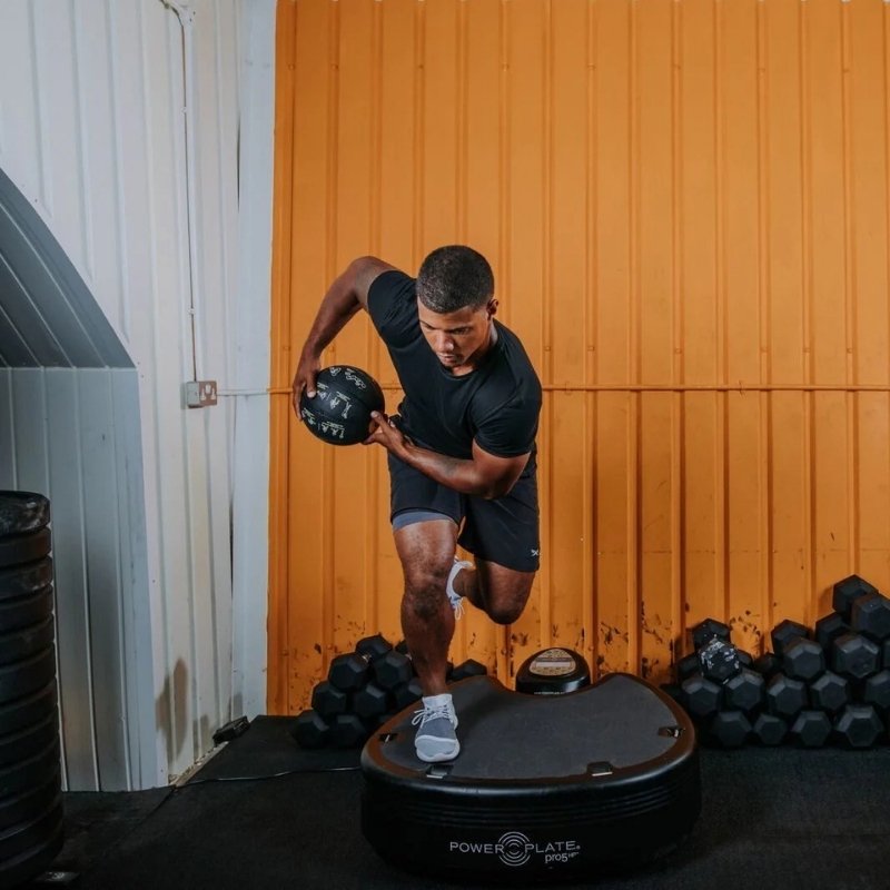 Person using a Power Plate machine in a gym setting with orange walls and dumbbells.