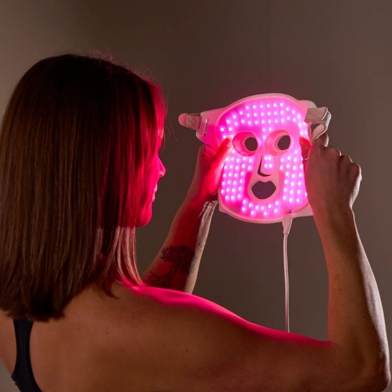 A woman holds a white Floe LED light therapy face mask glowing with pink light against a dark background.