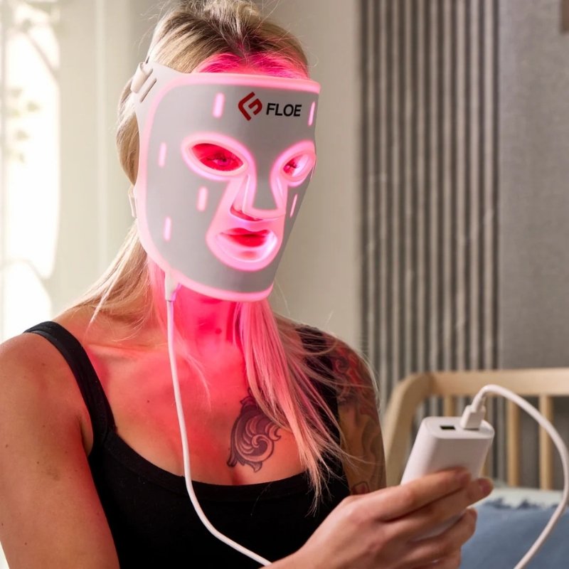 Person wearing a Floe LED face mask with visible branding, holding a device.