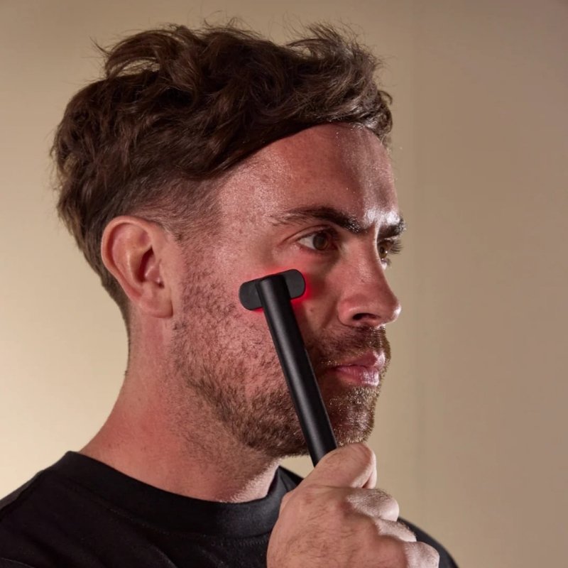 A man holding a black handheld Floe face wand device against his cheek, with a red light glowing from the tip, set against a plain beige background.