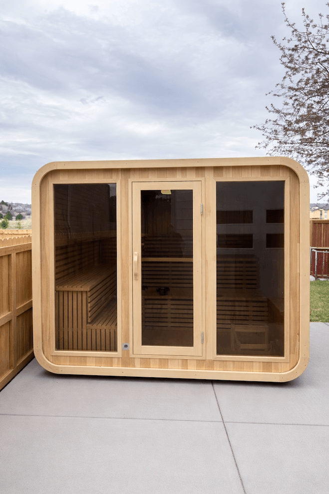 Wooden sauna cabin with glass doors on a concrete surface under a cloudy sky.